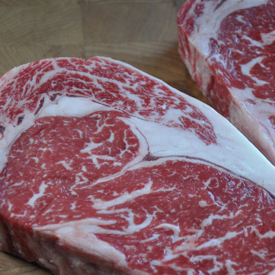 Two raw marbled beef steaks, with the front cut showing a thick layer of white fat over deep red meat, photographed on a wooden surface.